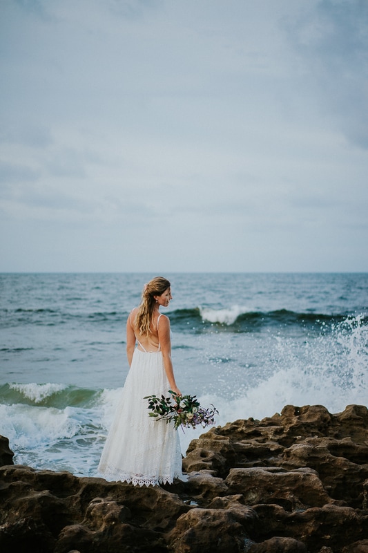 Coral Cove beach elopement bride in white lace beach wedding dress holding bouquet while wave crashes into rocks