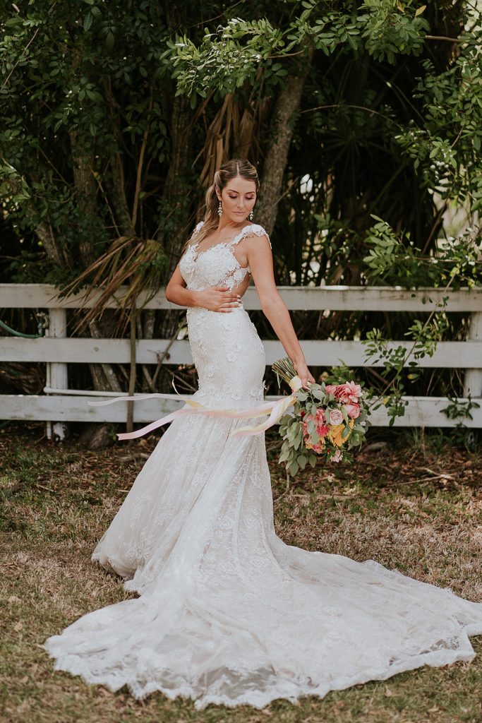 Bridal portraits in front of white fence with wind blowing the ribbons of her bouquet at Pink Lemonade photoshoot barn wedding at Twisted Oak Farm in Vero Beach FL