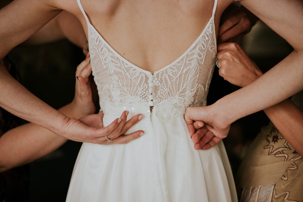 Close-up of the back of the wedding dress with lace bodice and buttons as friends help the bride into the dress