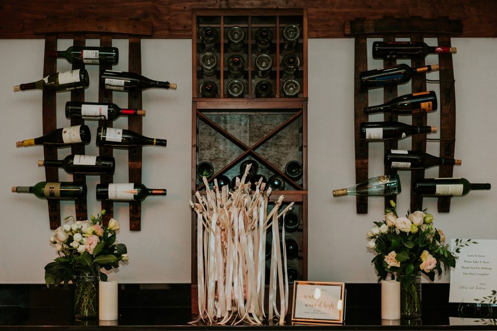 Pink and white wedding wands in a jar beneath wall display of wine bottles