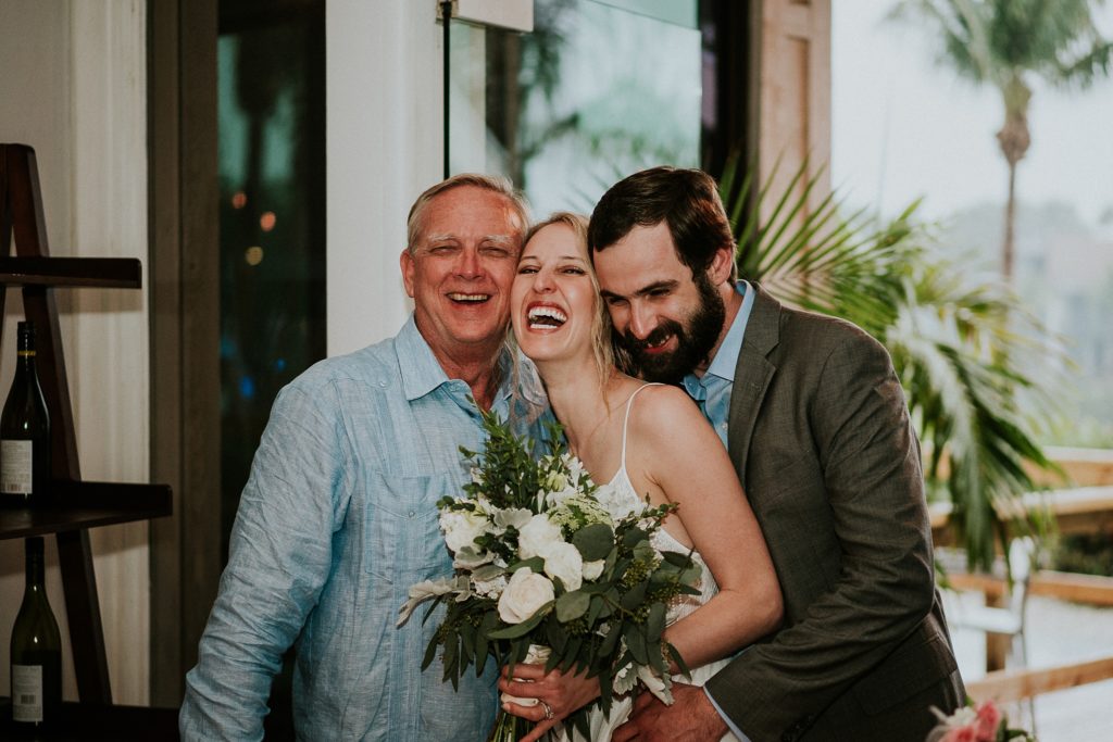 Father of the groom, the bride, and the groom hold each other and laugh