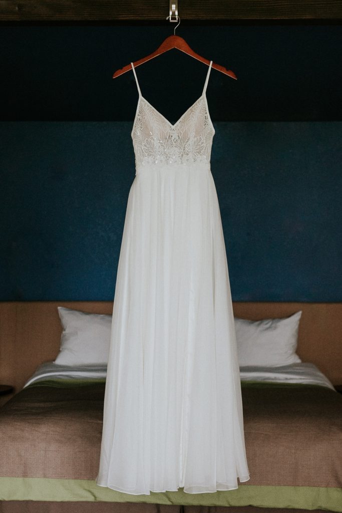 White A-line wedding dress with lace bodice hanging from a wood hanger above hotel bed with blue walls