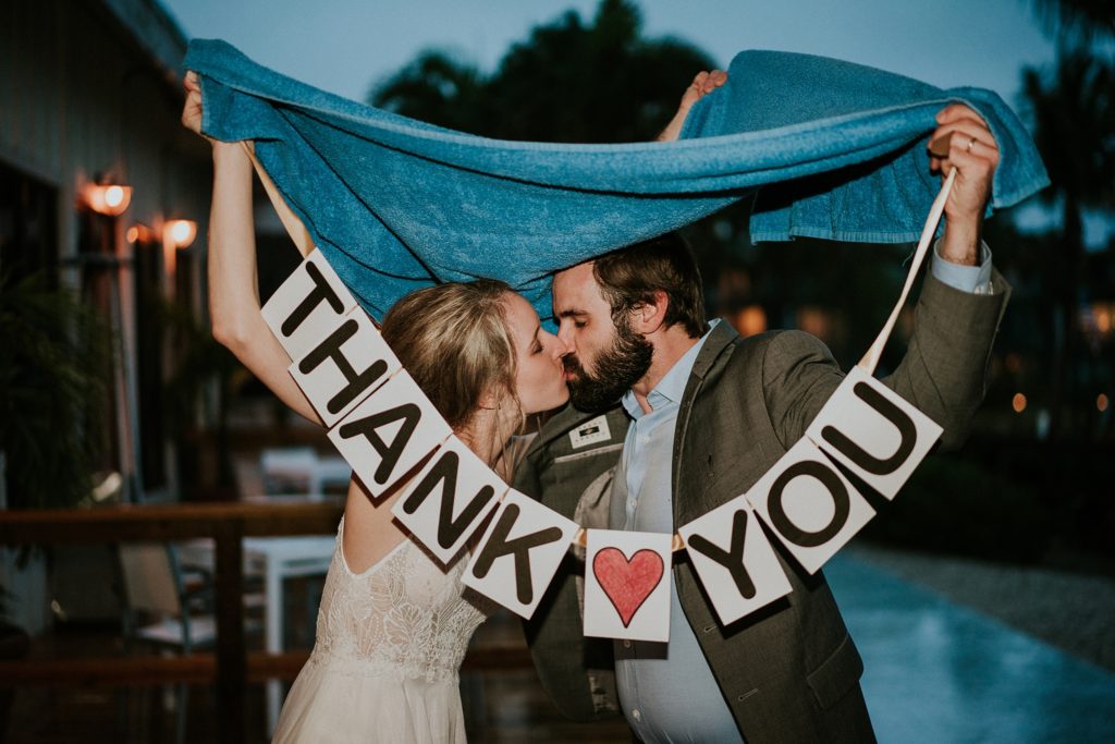 Bride and Groom kiss under a blue towel while holding Thank You sign outside on rainy wedding day