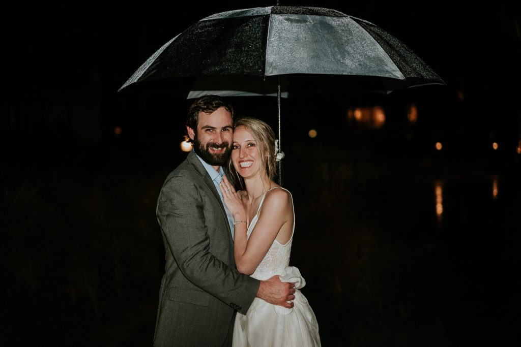 Wedding couple smiling at camera while holding an umbrella during rainy wedding day at night