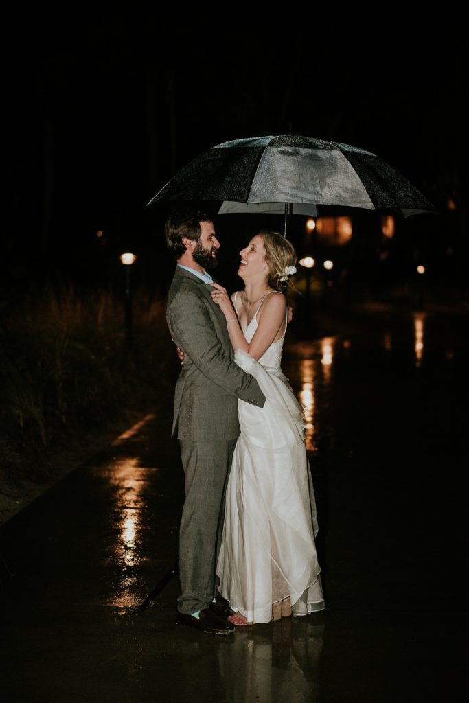 Bride and groom embrace under an umbrella for rainy wedding day photos outside at night