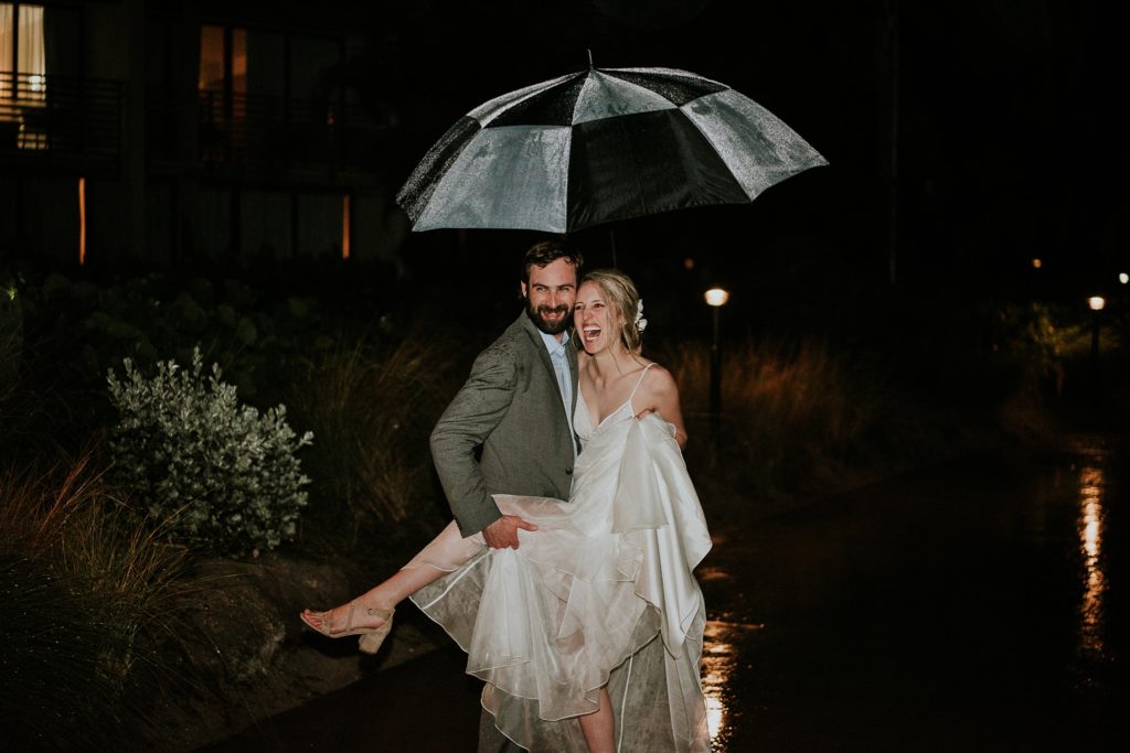 Groom holds bride's leg while they hold an umbrella for rainy wedding day photos at night