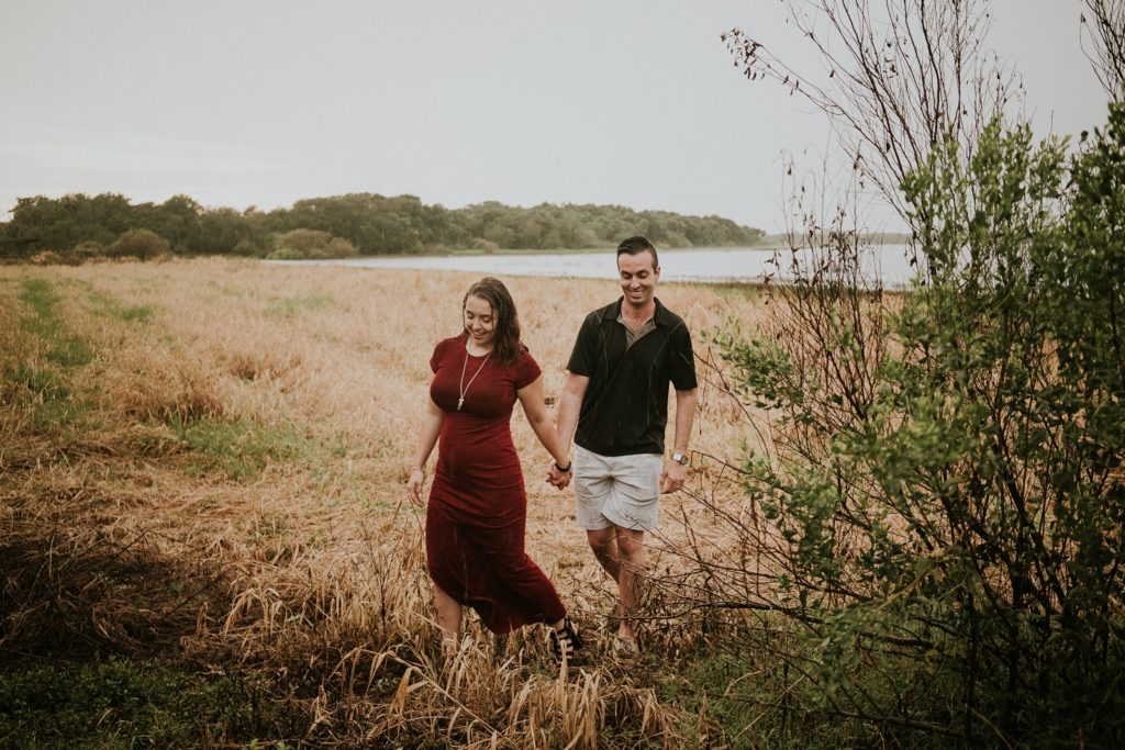 Couple holds hands and walks through golden field at Myakka river state park for engagement photos