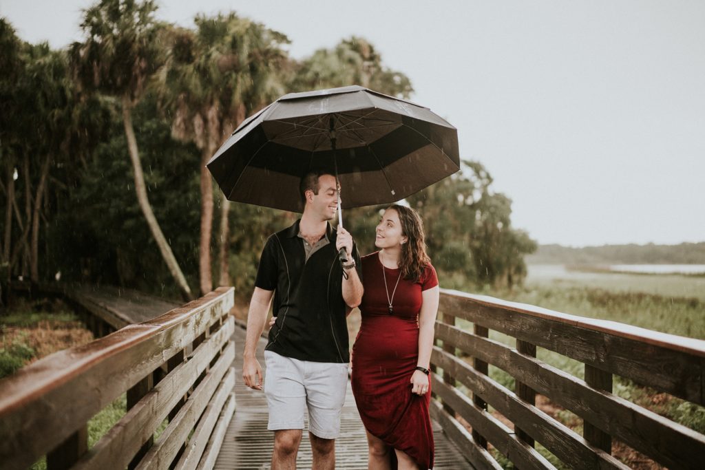 Myakka River State Park rainy engagement photos with Sarasota wedding couple looking lovingly at each other and holding umbrella on birdwalk at sunset