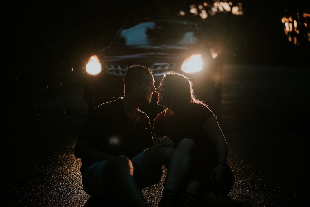 Headlights of a car illuminate the silhouette of a couple sitting on the ground and touching noses