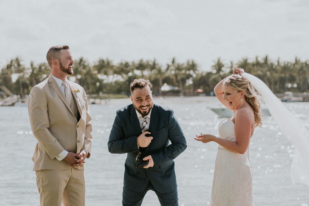 Groom in khaki suit and bride laugh as wind blows off her wedding veil during ceremony