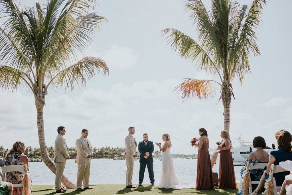 Bride reads vows to groom on phone on Sailfish Marina wedding lawn with two palm trees on the water in Singer Island