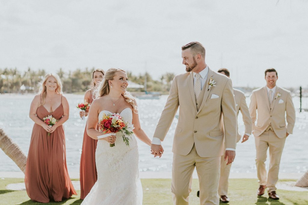 Bride and groom look at each other holding hands with wedding party behind in rust dresses and khaki suits