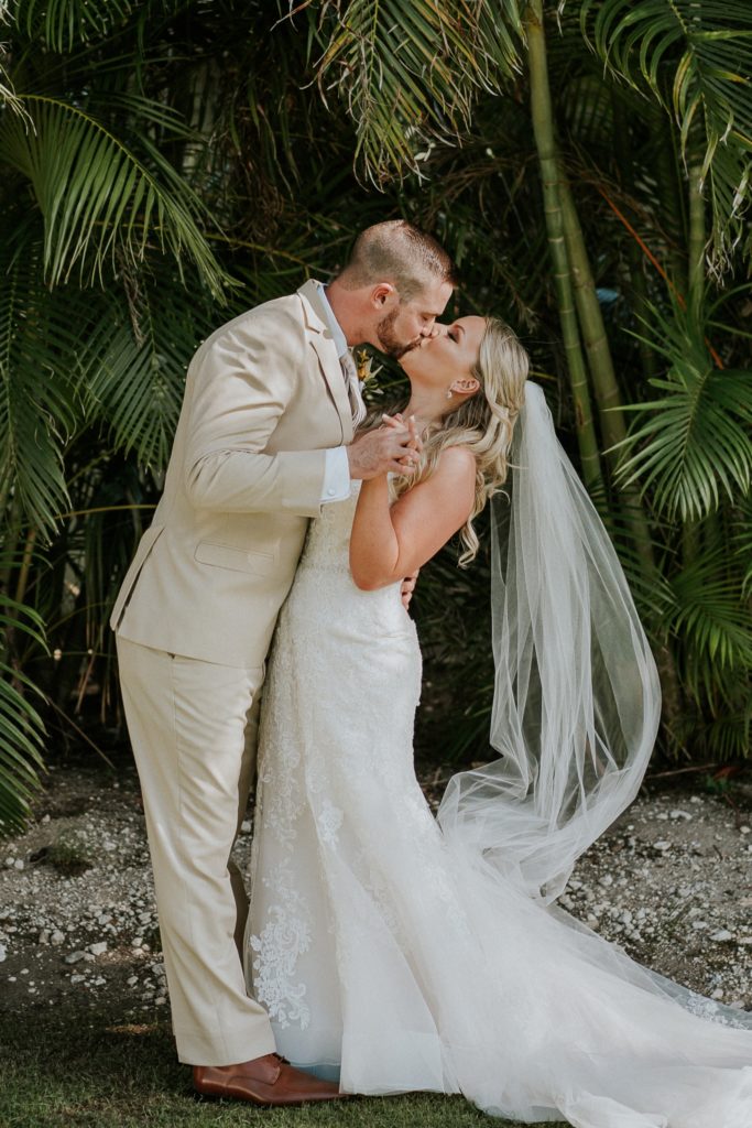 Bride and groom dip kiss with long chapel veil in front of green palm fronds and bamboo at Sailfish Marina Singer Island FL wedding venue