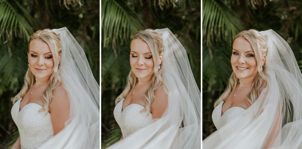 Bride looking down and smiling wearing long wedding veil flowing in front of her with green palm fronds behind