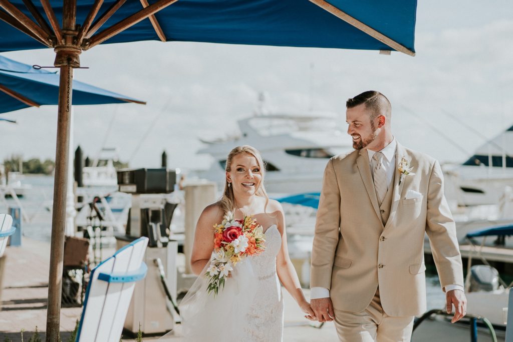 FL destination wedding couple hold hands walking Sailfish Marina Resort smiling under blue beach umbrella on Singer Island