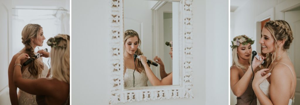 Bridesmaid helps bride curl hair in front of mirror in white bathroom