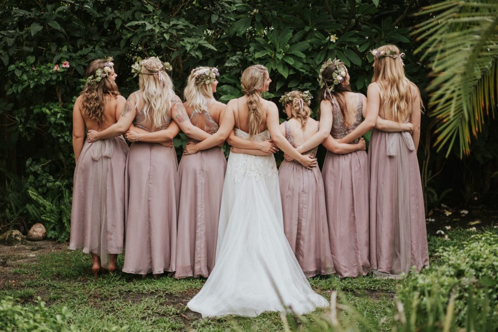 Bride and bridesmaids in lilac dresses link arms with backs to camera