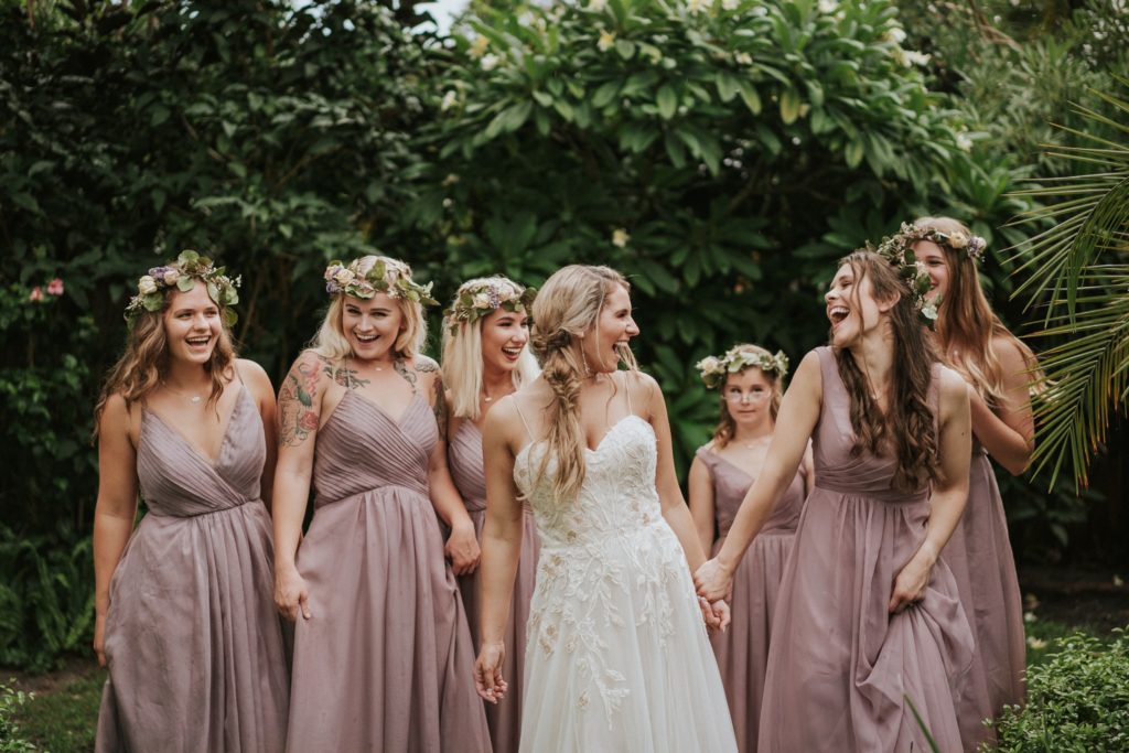 Bride and bridesmaids in lilac dresses and flower crowns walk towards camera and laugh