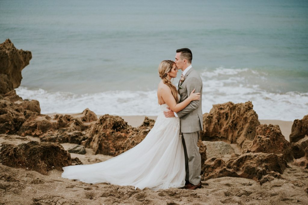 Groom kisses bride's forehead as they embrace on rocky beach at House of Refuge elopement with ocean behind them