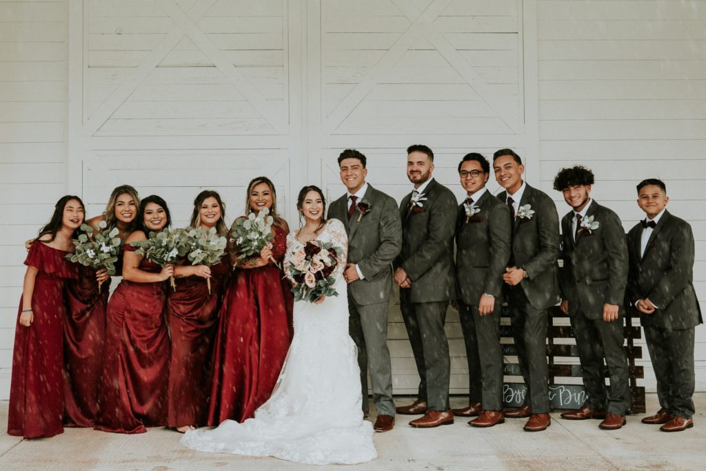Bridesmaids wearing red dresses and groomsmen in grey suits stand next to bride and groom in front of white barn doors in the rain
