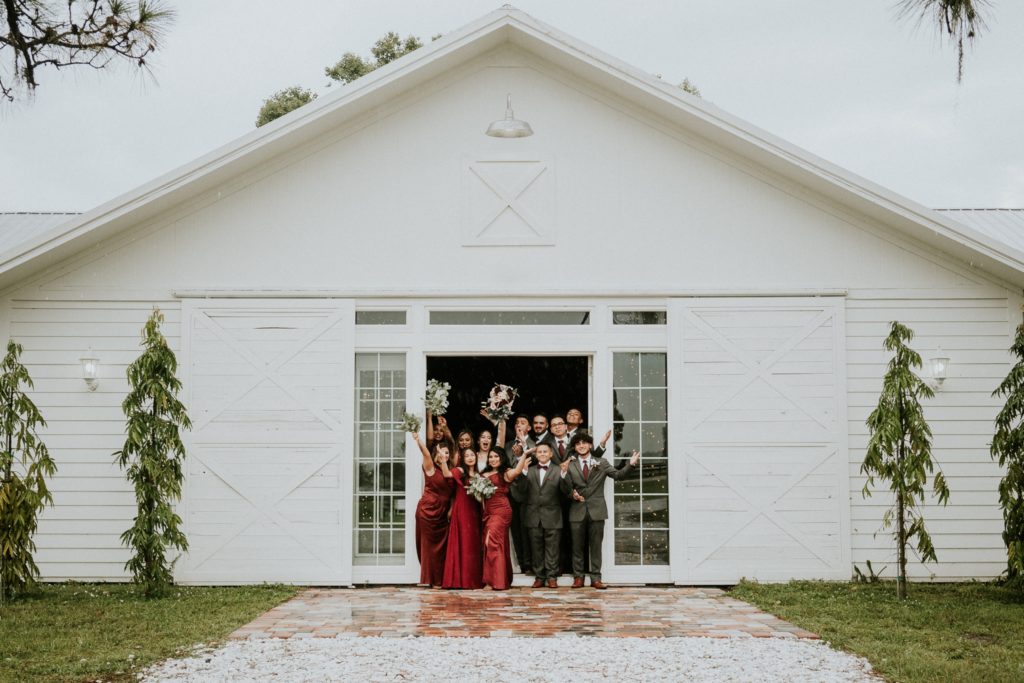 Bridesmaids in dark red dresses and groomsmen cheer with bride and groom in front of french doors at Ever After Farms Ranch white Wedding Barn in Indiantown FL