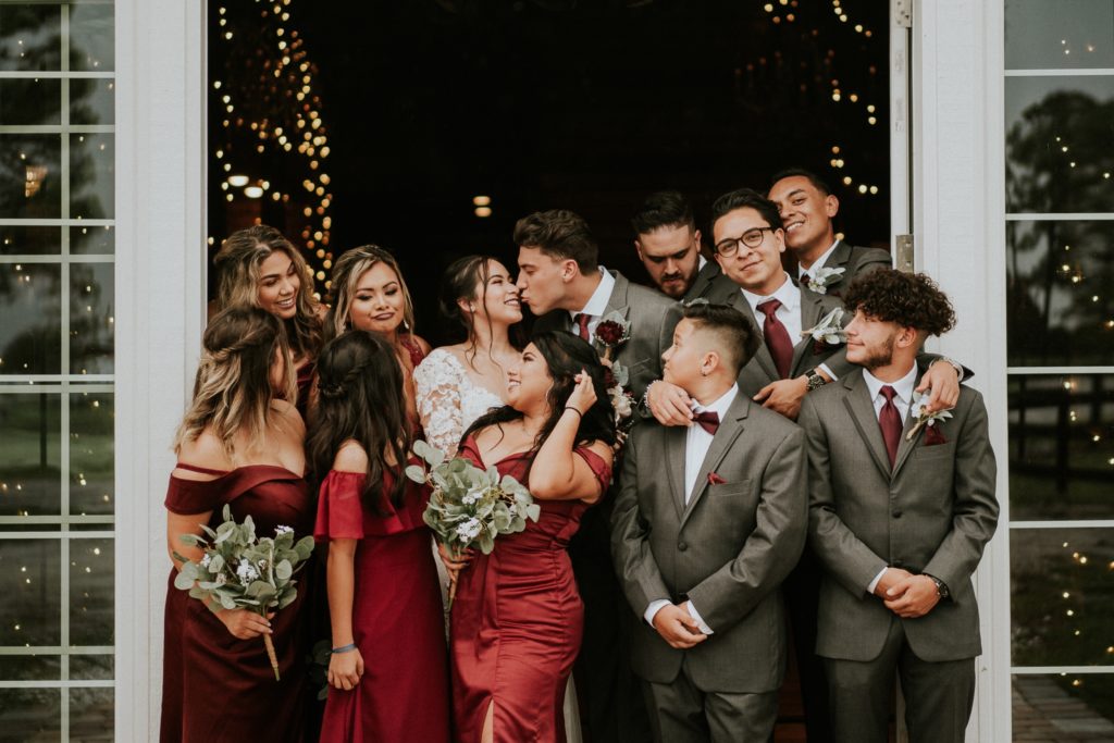 Bridesmaids wearing dark red dresses and groomsmen wearing grey suits and dark red ties look at bride and groom as they kiss at Ever After Farms Ranch Wedding Barn in Indiantown