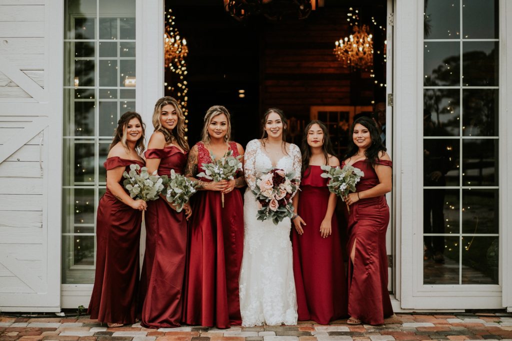 Bridesmaids wearing dark red dresses with bride in front of white french doors with fairy lights behind them at Ever After Farms Ranch in Indiantown