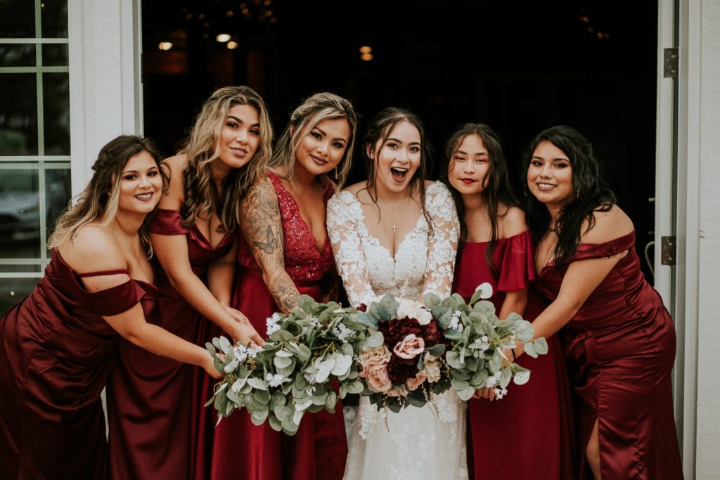 Bride in white lace floral dress with bridesmaids wearing red dresses hold their bouquets out and smile