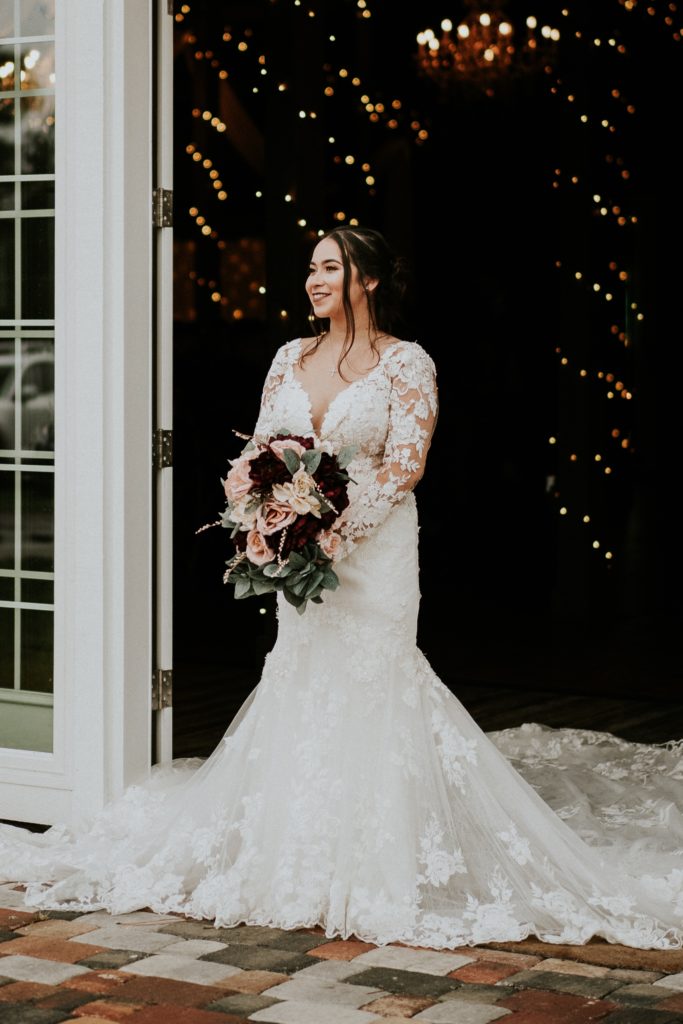 Bride stands by white french doors on cobblestones and looks away from camera holding fake flower bouquet and wearing Martina Liana designer wedding gown