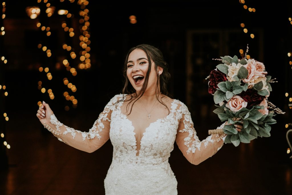 Bride holds pink and red flower bouquet in air and cheers with fairy lights behind her