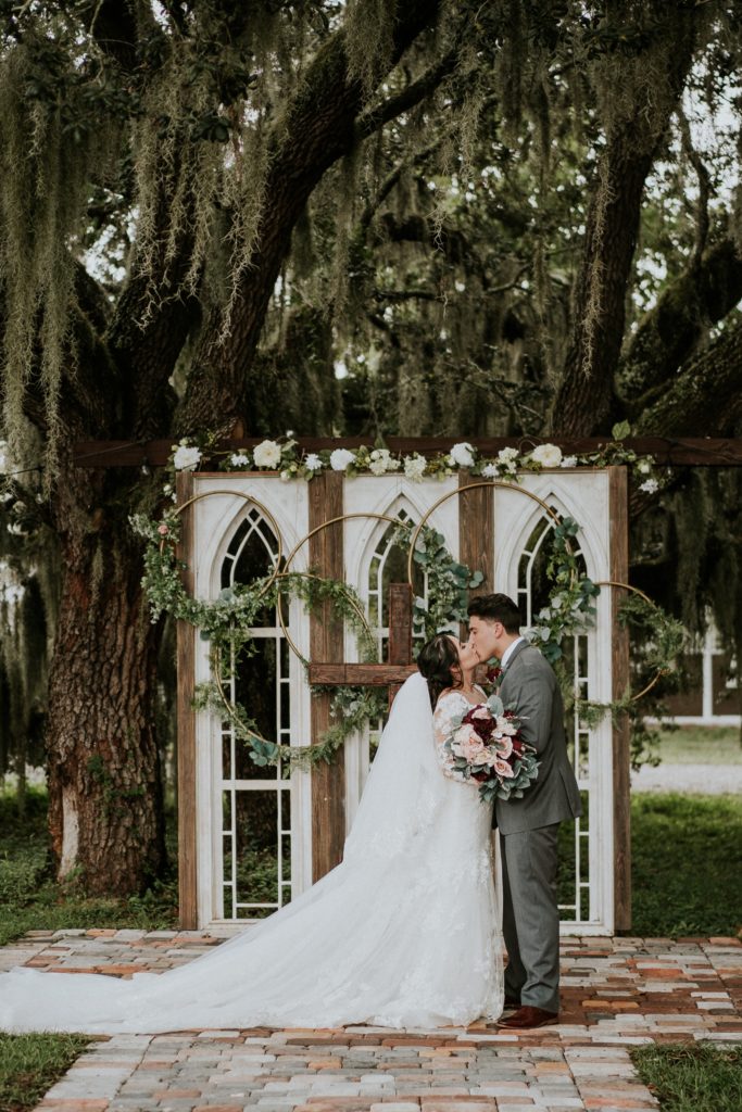 Bride and groom kiss in front of white gothic window wedding altar ceremony backdrop at Ever After Farms Ranch Barn in FL