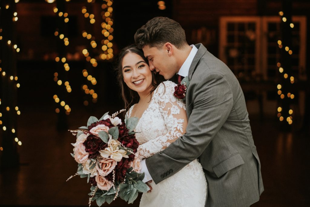 Bride smiles at camera holding pink and red rose bouquet as groom hugs her with fairy lights behind them