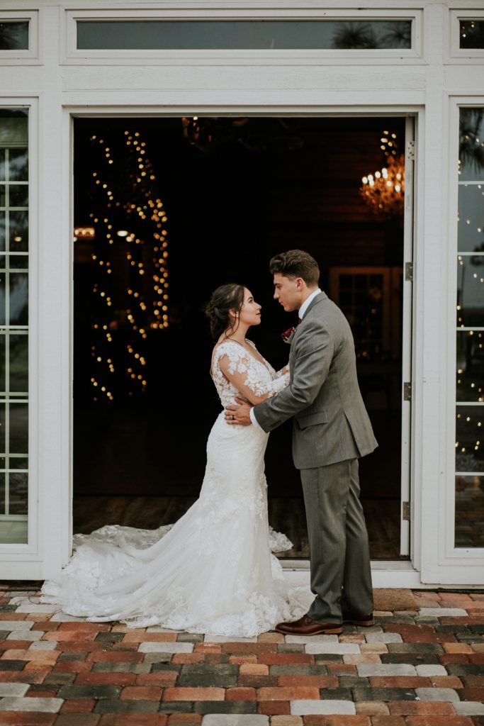 Bride and groom embrace in front of french doors of white barn wedding venue on cobblestones with fairy lights in background