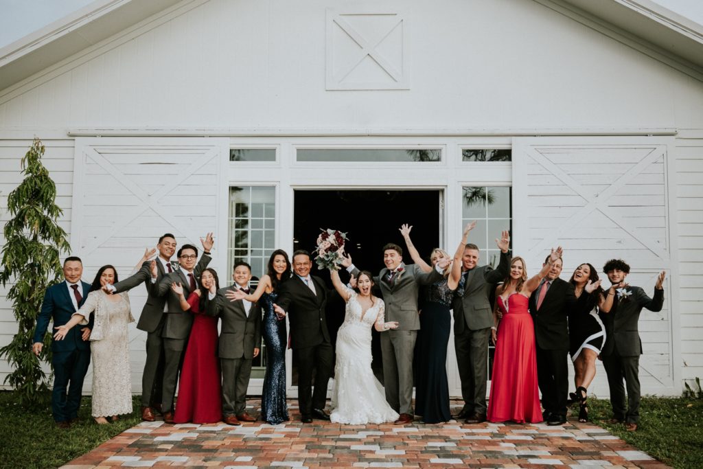 Family of bride and groom stand in front of white barn doors and throw hands in the air to cheer