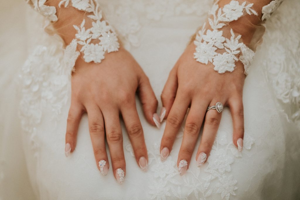 Close up of bride's hands wearing pear teardrop engagement ring and pink manicure with white flower and lace details