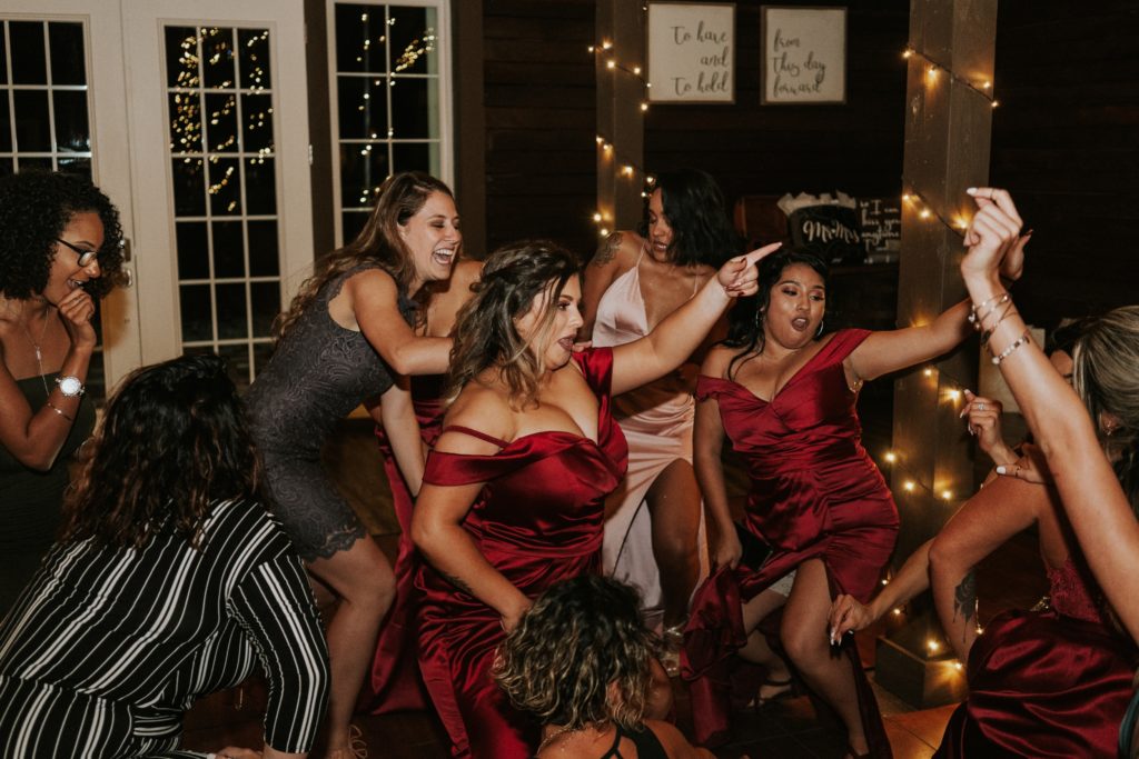 Bridesmaids in red dresses dance with guests at barn wedding reception with fairy lights
