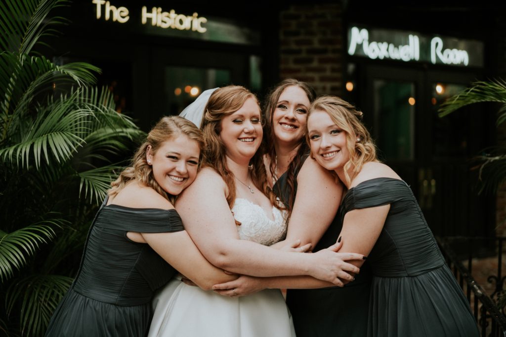 The bride and her sisters wearing green bridesmaid dresses group hug in the Historic Maxwell Room courtyard