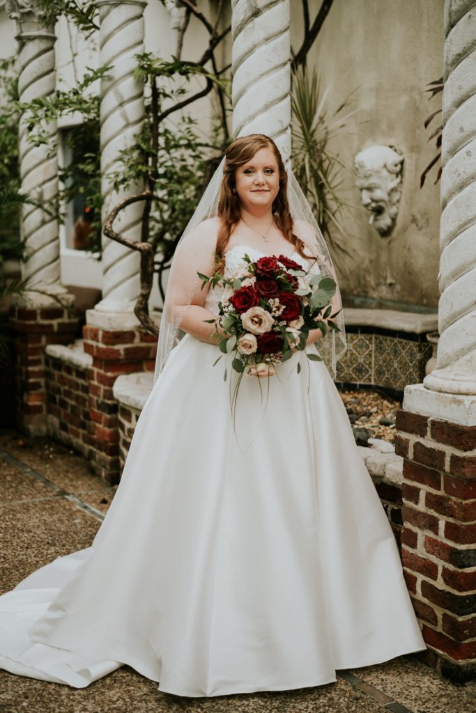 Redheaded Bride Allisha holds red and pink rose wedding bouquet smiling in front of pilllars and gargoyle grotesque at the Historic Maxwell Room courtyard