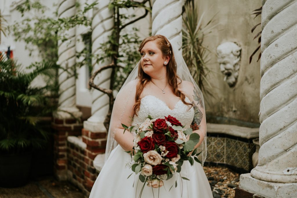 Redheaded Bride Allisha holds red and pink rose wedding bouquet and looks away in Historic Maxwell Room courtyard