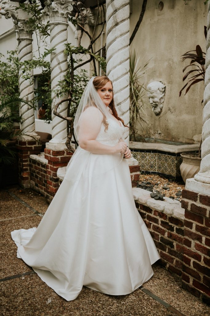 Bride wearing satin white wedding gown in front of brick ledge, gargoyle, and pillars at the Historic Maxwell Room courtyard