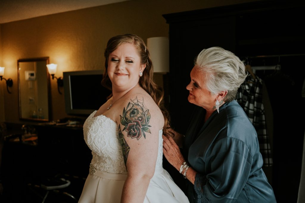 Grey-haired mother of the bride buttons her daughter's strapless wedding dress that shows off her arm planchette tattoo