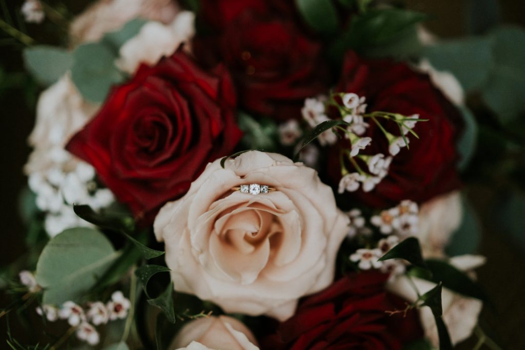 Gold diamond engagement ring nestled in petals of a pink rose in wedding bouquet with red roses and daisies