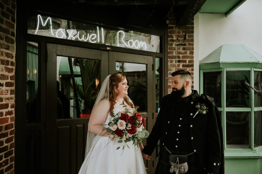 Bride Allisha and groom Stan hold hands and smile at each other in front of the Historic Maxwell Room neon sign doors