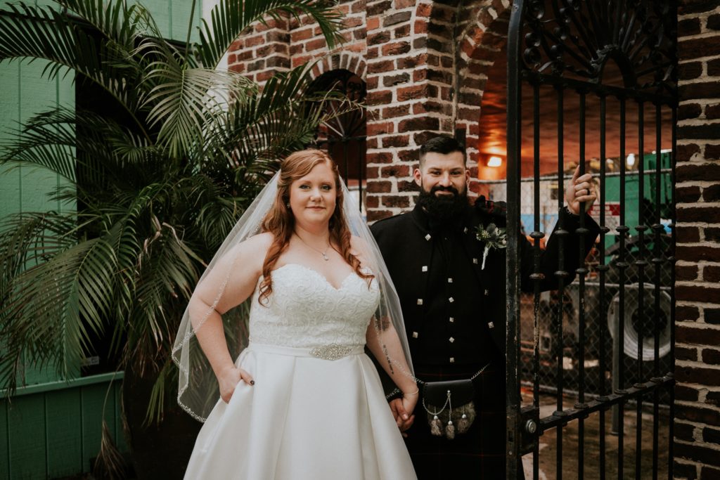 Bride places one hand in pocket of white wedding dress and holds hands with groom Stan who holds onto an iron fence in front of a brick wall at the Historic Maxwell Room courtyard