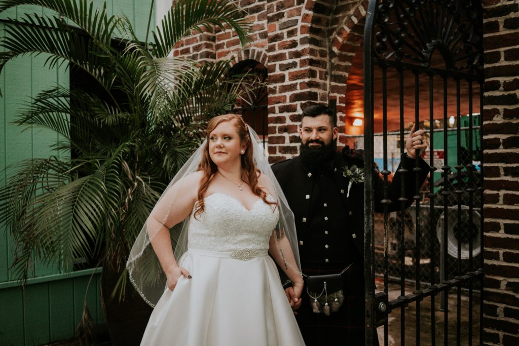 Bride places one hand in pocket of white wedding dress and holds hands with groom Stan who holds onto an iron fence in front of a brick wall at the Historic Maxwell Room courtyard