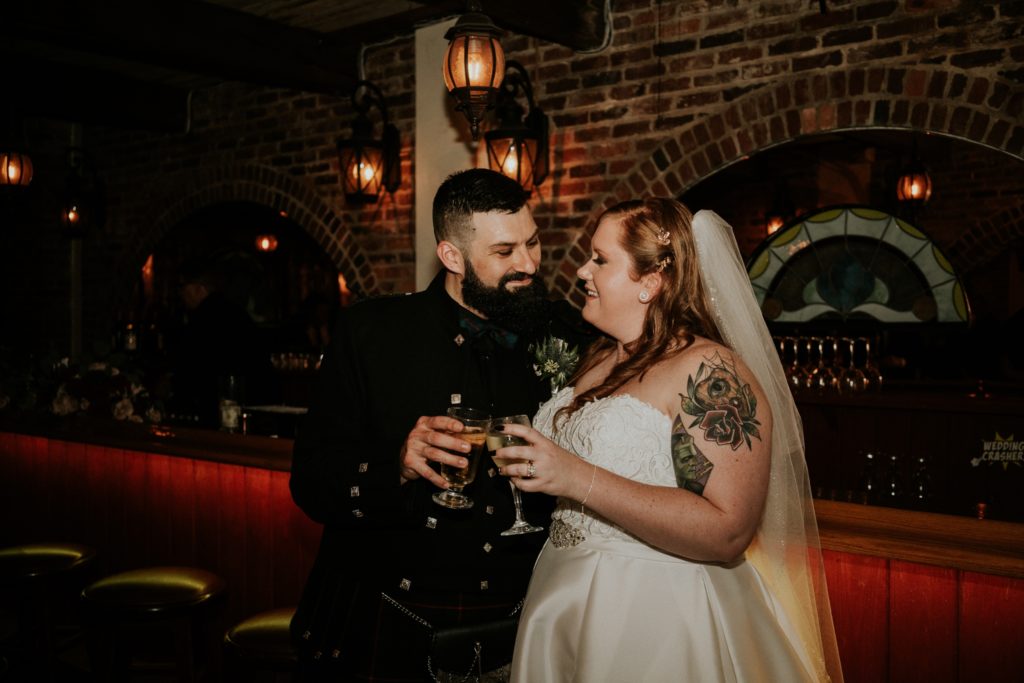 Groom Stan and bride Allisha smile and clink glasses at the Historic Maxwell Room wedding venue bar with brick walls and lit lanterns
