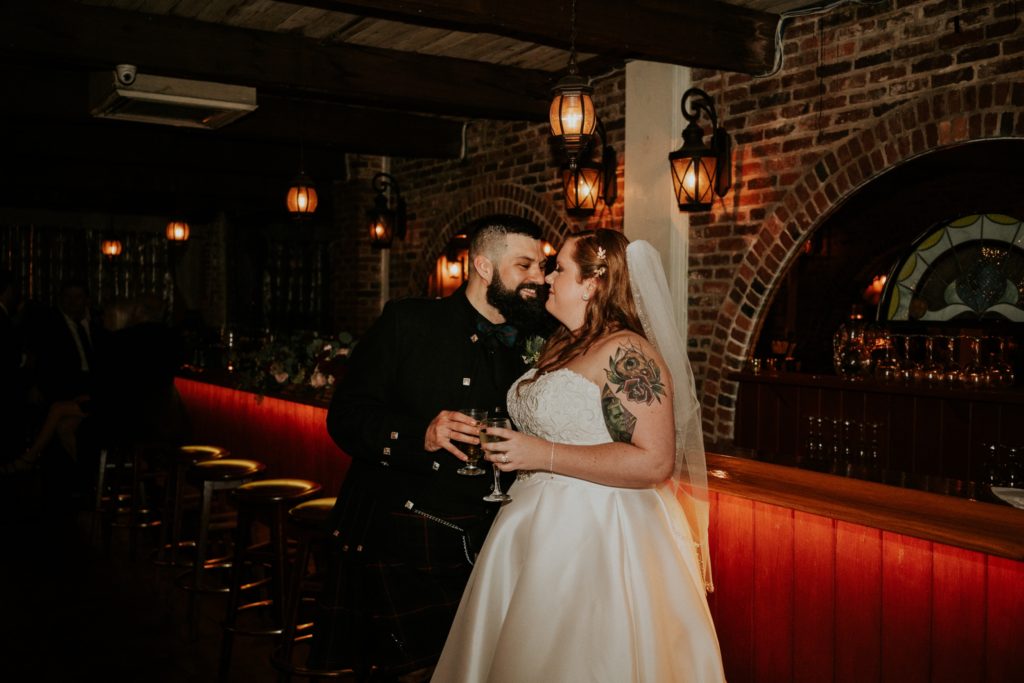 Groom and bride touch noses and clink glasses at the Historic Maxwell Room wedding venue bar with brick walls and lit lanterns