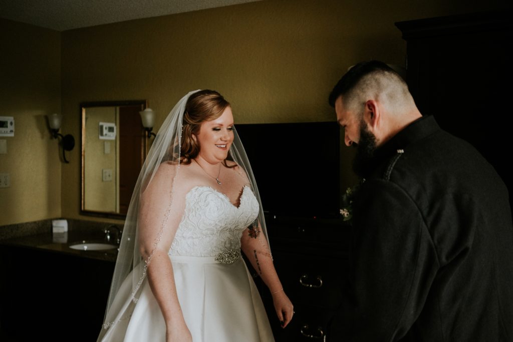 Groom sees his bride wearing her strapless white satin wedding dress and veil for first time in hotel room