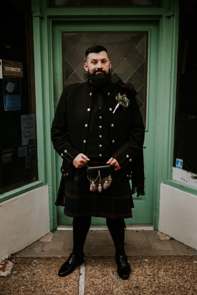 Groom Stan stands in front of bright green door wearing traditional Scottish wedding attire including kilt