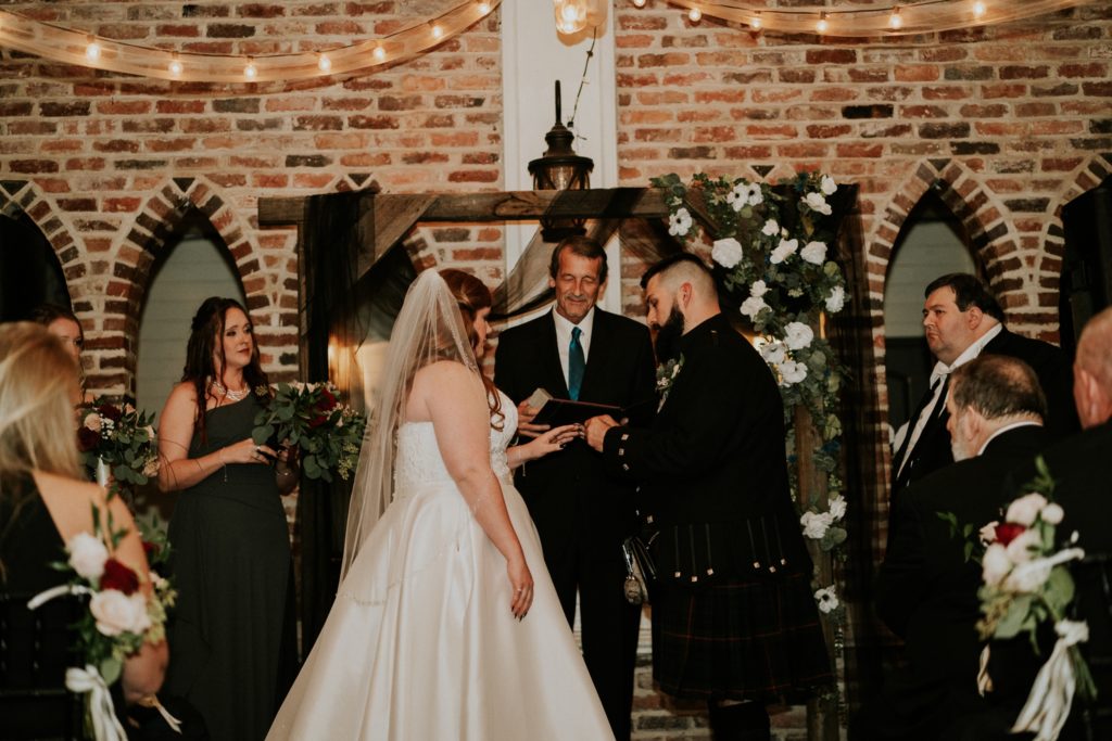 Bride and groom exchange wedding rings during indoor ceremony under wood arch with black chiffon drapery and white roses at the Historic Maxwell Room
